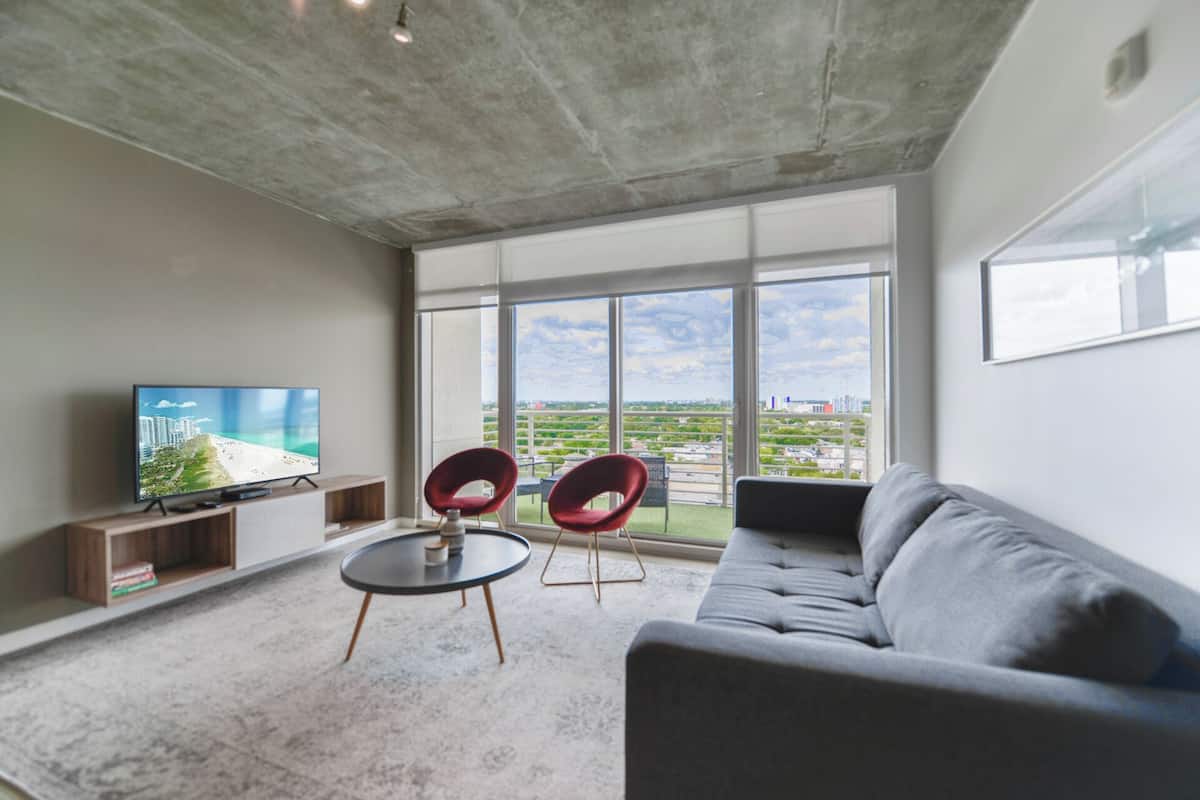 A modern living room with a grey sofa, two red chairs, a small round table, and a TV on a wooden stand. Large windows offer a view of a cityscape and greenery.