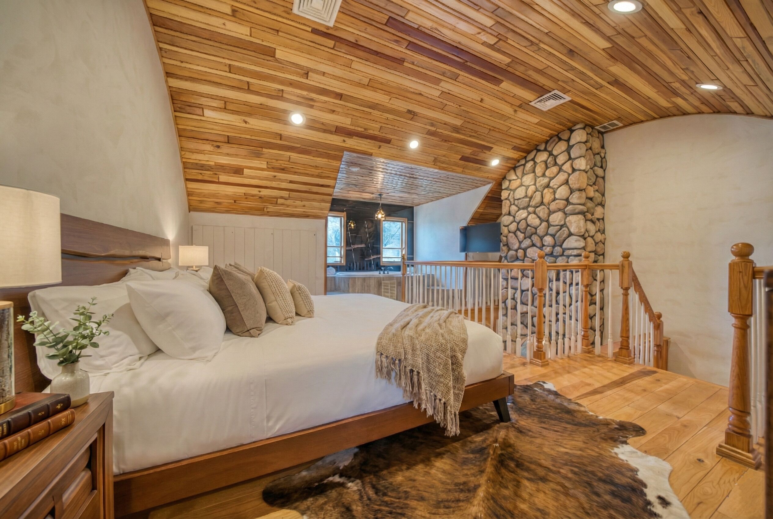 A bedroom with a wooden ceiling, a stone wall, a bed with white bedding, a wooden floor, and a view of a balcony.