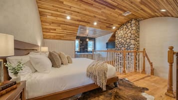A bedroom with a wooden ceiling, a stone wall, a bed with white bedding, a wooden floor, and a view of a balcony.