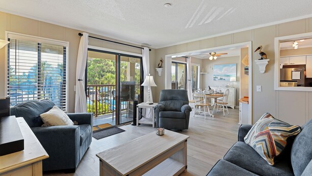 A modern living room with a sofa, armchair, and coffee table. There is a dining area with a table and chairs visible through an open door. - 2 BR Updated Admirals Row at HH Beach and Tennis Bikes & Beach Gear included (Hilton Head Island)