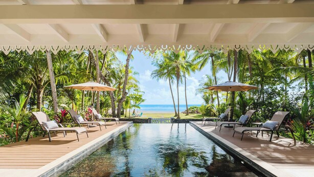 Un espace aménagé au bord de la piscine, avec des chaises longues et des parasols, entouré de palmiers et offrant une vue sur l’océan.