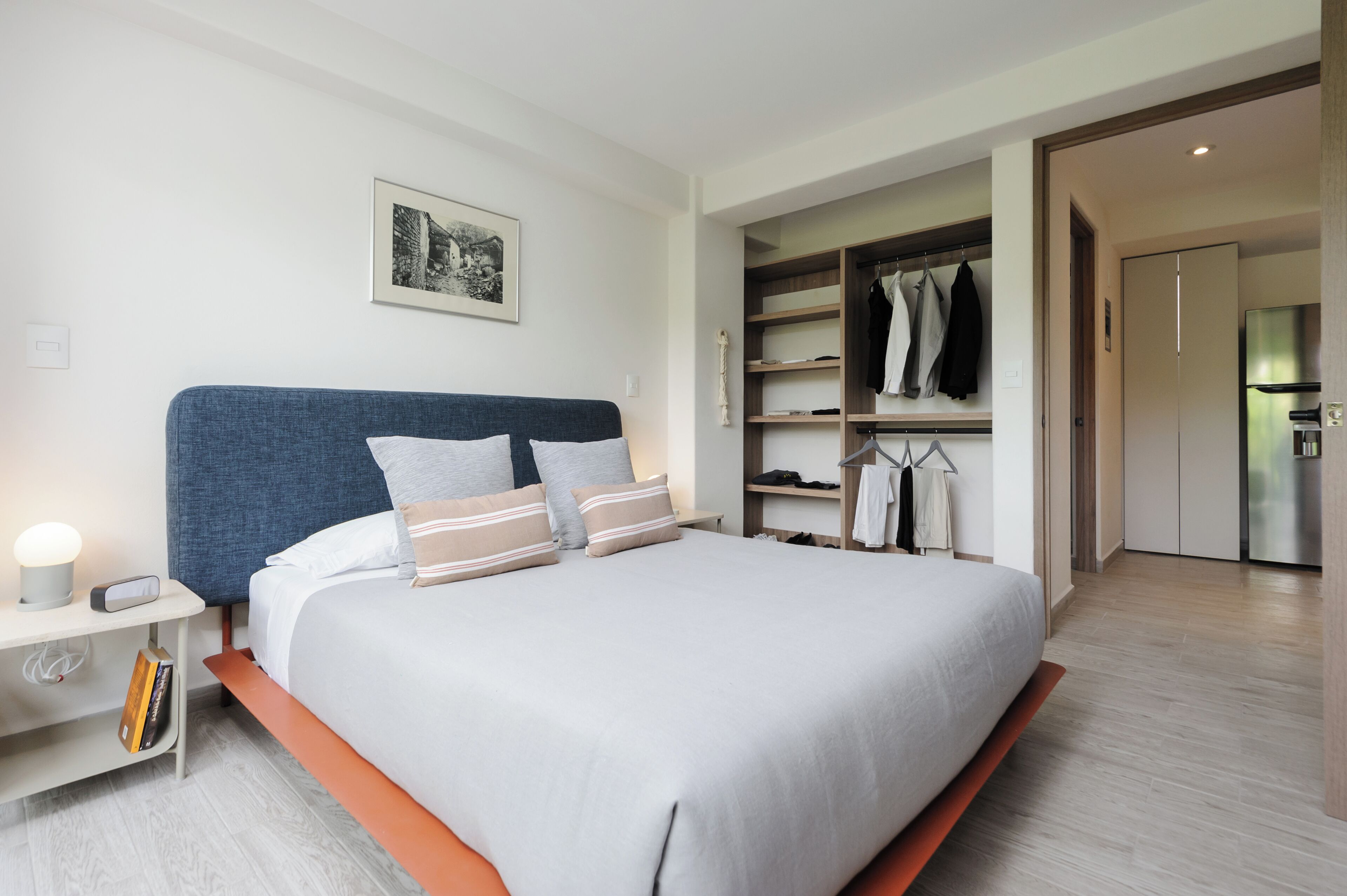 Quarto moderno com uma cama grande, mesa de cabeceira com abajur, guarda-roupa e um quadro na parede.
