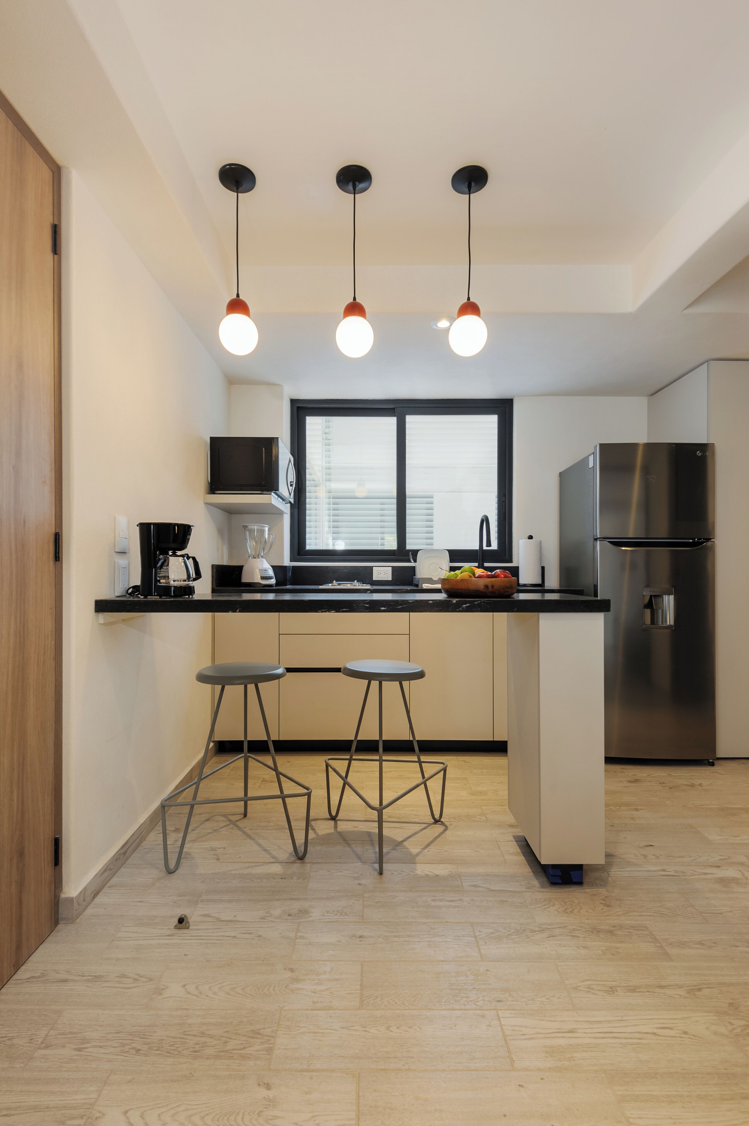 Una cucina moderna con un piano di lavoro nero, un frigorifero in acciaio inossidabile, un forno a microonde e una macchina da caffè. Sono presenti due sgabelli e una finestra con persiane.