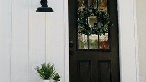 Un bâtiment blanc avec une porte noire, un petit porche avec une table et des plantes en pot, et une boîte aux lettres noire fixée au mur.