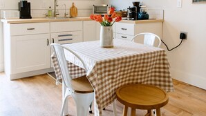 Une cuisine moderne avec des meubles blancs, des étagères en bois et un coin repas agrémenté d’une nappe à carreaux.
