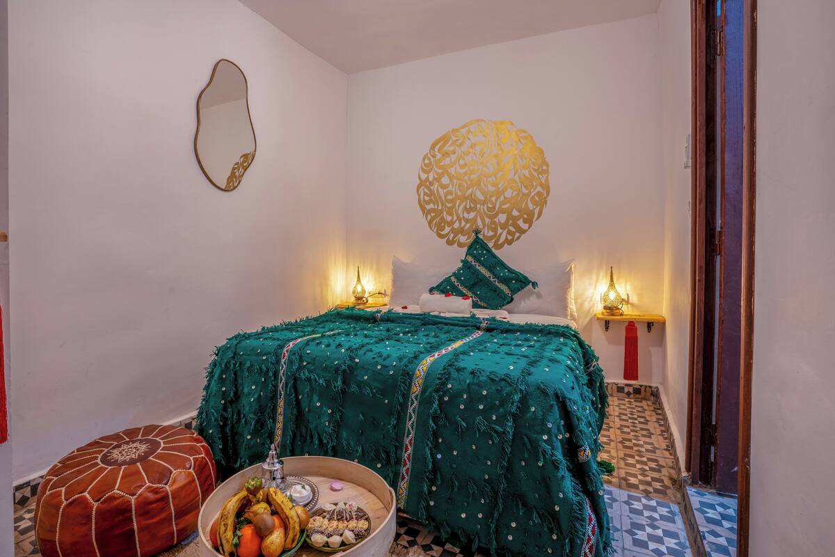 A room with a bed, a wall-mounted mirror, a decorative wall piece, a small table with fruits, and a patterned floor.
