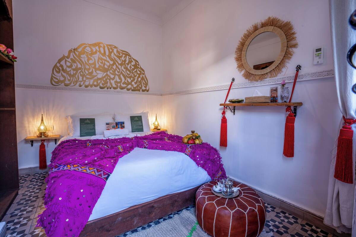A room with a bed, a wall-mounted mirror, a shelf with decorative items, and a patterned floor.