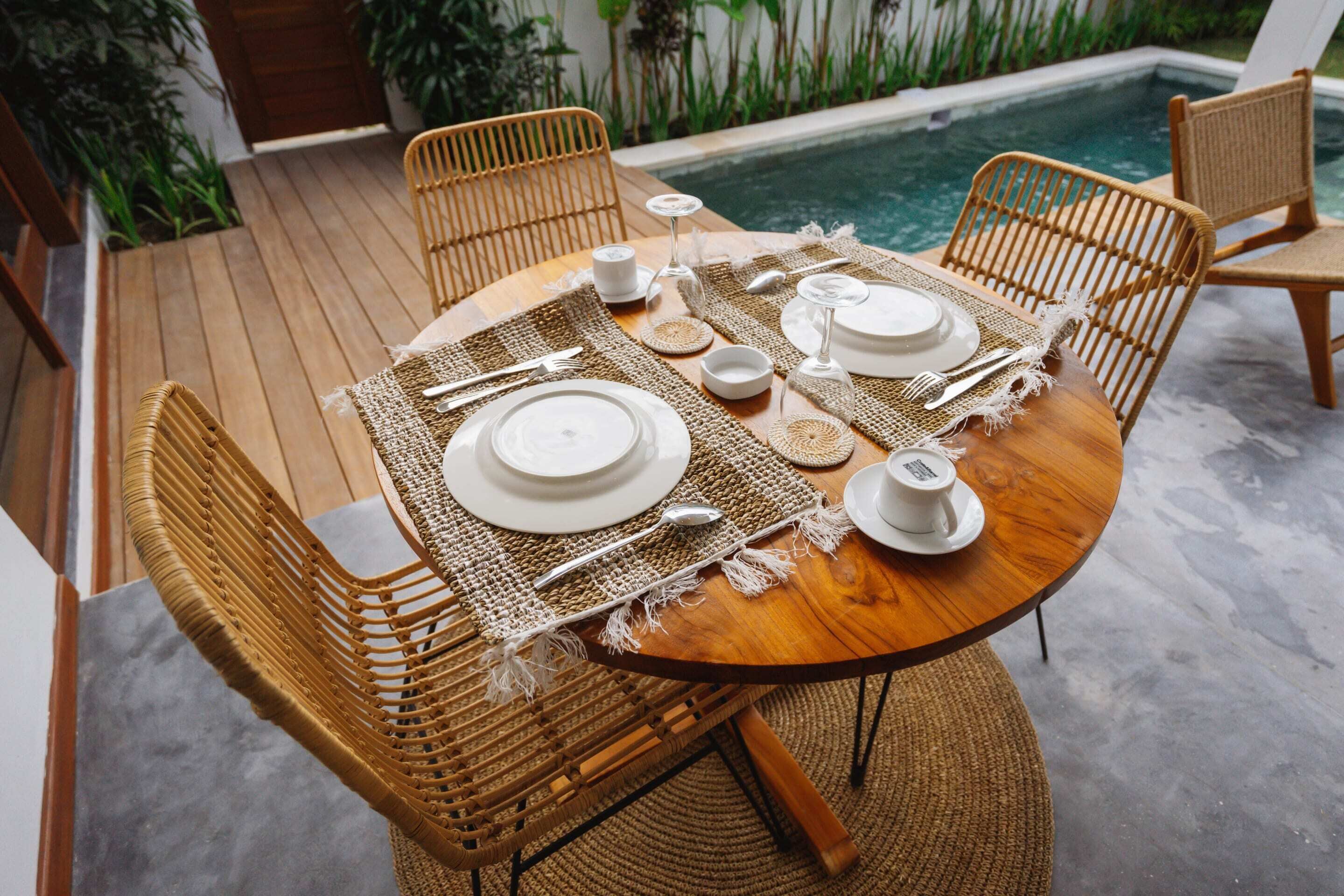 Una mesa de comedor de madera dispuesta con platos blancos, cubiertos y copas, rodeada de sillas de mimbre, con una piscina al fondo.