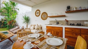A modern kitchen with wooden cabinets, a dining area with a round table set for four, and a cozy seating area with a sofa and cushions. - Villa Karina (Tabanan)