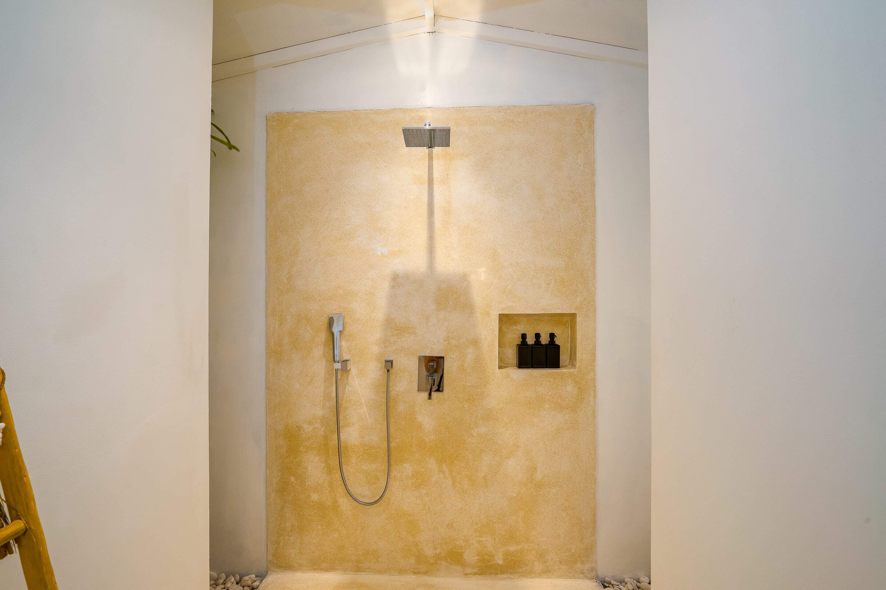 A shower with a handheld showerhead and a wall-mounted faucet, a small shelf with bottles, and a ladder in the corner.
