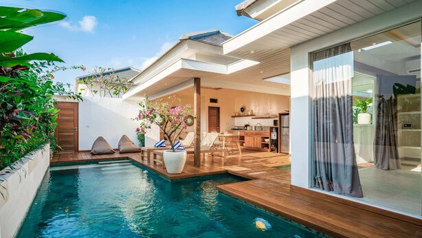 Ein moderner Poolbereich im Freien mit Holzterrasse, Liegestühlen und einem durch eine offene Tür einsehbarer Küchenbereich.