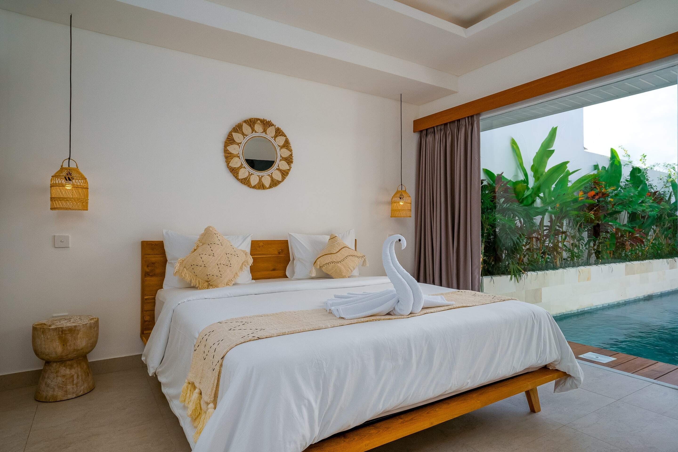 Une chambre avec un grand lit, une tête de lit en bois, un miroir rond et une vue sur une piscine.
