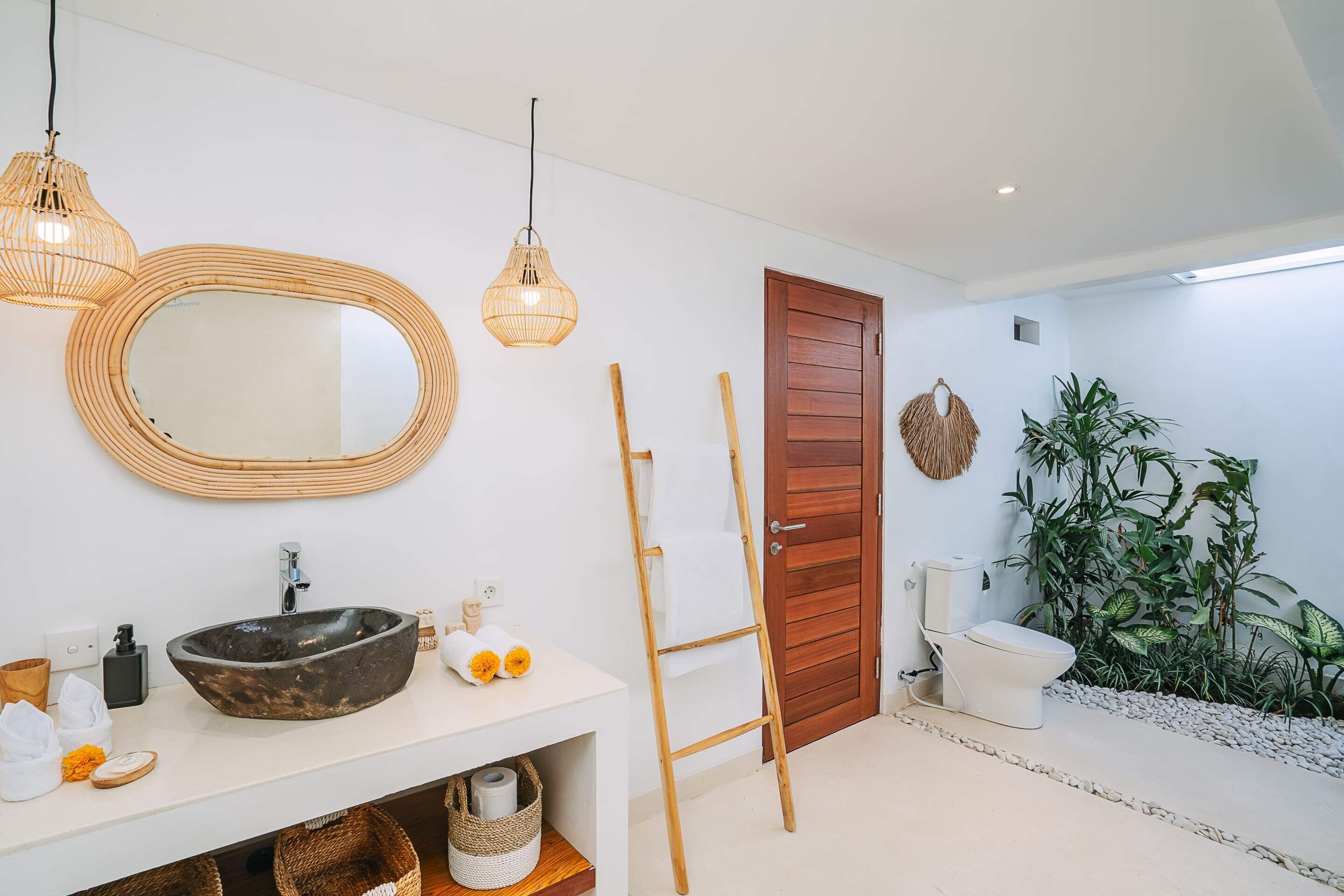 A modern bathroom with a large oval sink, two hanging wicker light fixtures, a wooden ladder, a wooden door, and a toilet.