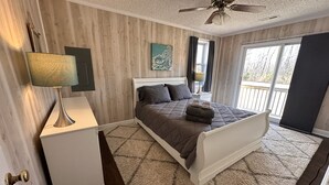 A bedroom with a bed, a nightstand, a lamp, a ceiling fan, and a window with curtains. - Carrick Bend -Balcony View of ICW- Dog Friendly! (Coinjock)