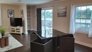 A living room with a flat-screen TV, a glass-top dining table, and a view of a lake through large windows. - Carrick Bend -Balcony View of ICW- Dog Friendly! (Coinjock)
