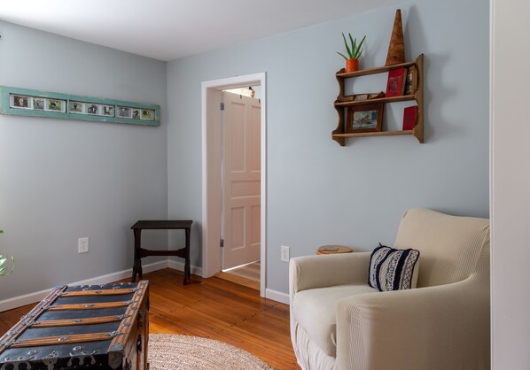 A living room with a sofa, a small table, a wooden bench, a wall-mounted shelf with books, and a door leading to another room. - 1900’s farmhouse with old country charm. Six miles to Lake Wallenpaupack (Lake Ariel)