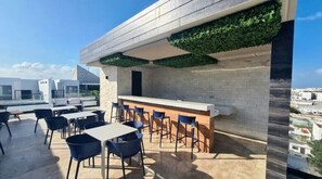 Relax on the outdoor terrace with a bar, chairs, and tables, surrounded by lush greenery and a city view.