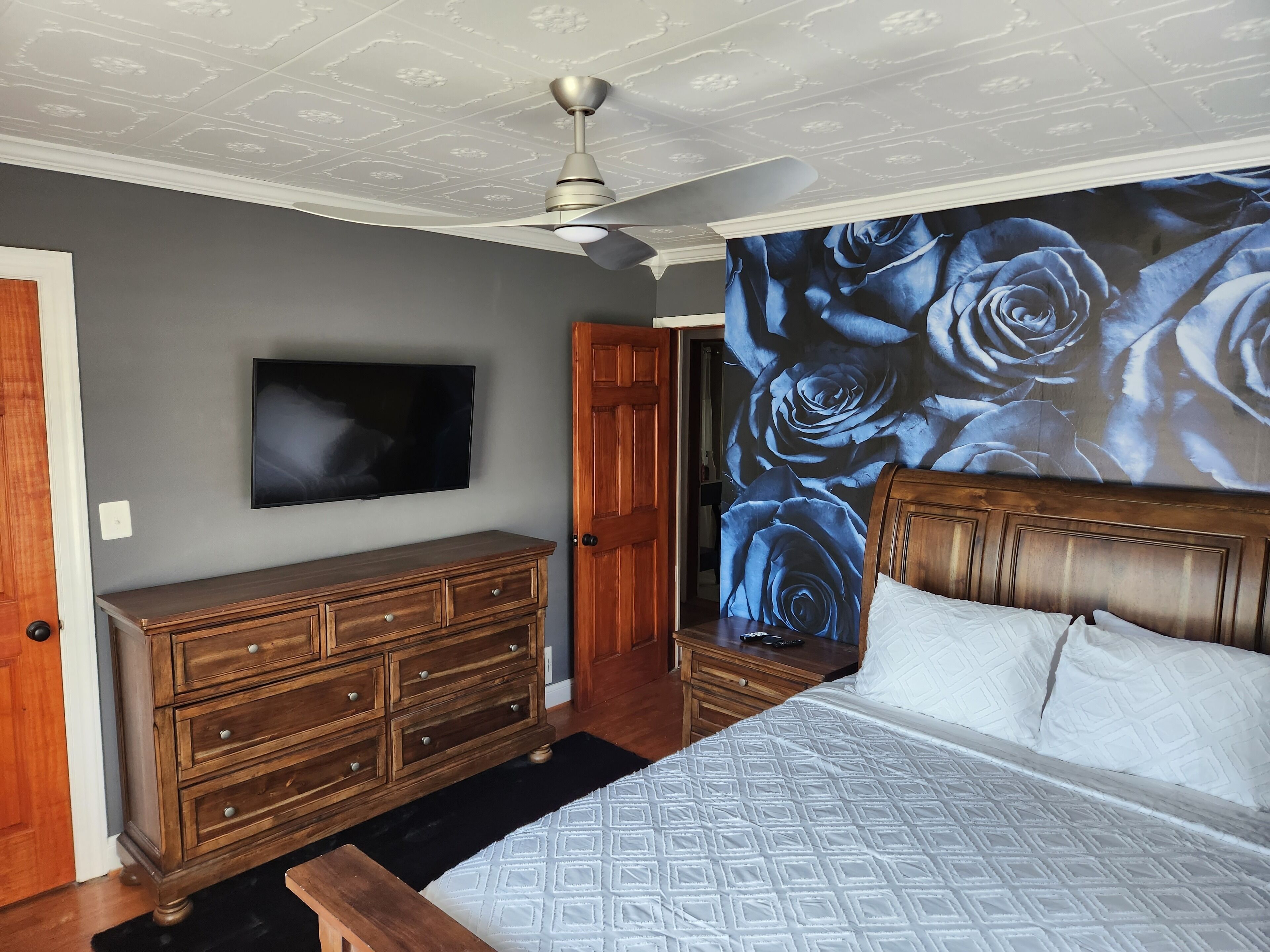Un dormitorio con cama, cómoda de madera, ventilador de techo, televisor y una pared con un mural de rosa azul.