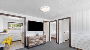 A modern, well-lit room with a wooden dresser, a TV mounted on the wall, and a window providing a view of the outdoors. - Relax at Lazy Duck Inn #18: a 2BR suite with kitchenette, just 1 minute to Yellowstone. (West Yellowstone)