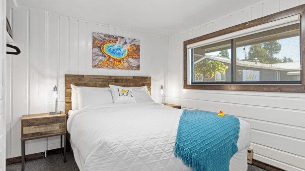 A bedroom with a wooden headboard, white bedding, a blue throw blanket, a nightstand with a lamp, and a window with a view of the outside. - Relax at Lazy Duck Inn #18: a 2BR suite with kitchenette, just 1 minute to Yellowstone. (West Yellowstone)