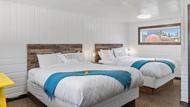 A hotel room with two beds, white bedding, a wooden headboard, a yellow side table, and a window with a view of a building. - Stay at Lazy Duck Inn #20 Double Queens, just 1 minute to Yellowstone! (West Yellowstone)