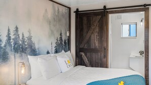 A bedroom with a bed, a nightstand, a lamp, a wall mural, a sliding barn door, a window, and a bathroom visible through the door. - Cozy Lazy Duck Inn #22: 1 min to Yellowstone, 2 full beds, kitchen, sleeps 4! (West Yellowstone)