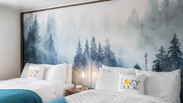 A hotel room with two beds, each with a pillow featuring the hotel's logo. A large forest mural adorns the wall behind the beds. - Cozy Lazy Duck Inn #22: 1 min to Yellowstone, 2 full beds, kitchen, sleeps 4! (West Yellowstone)