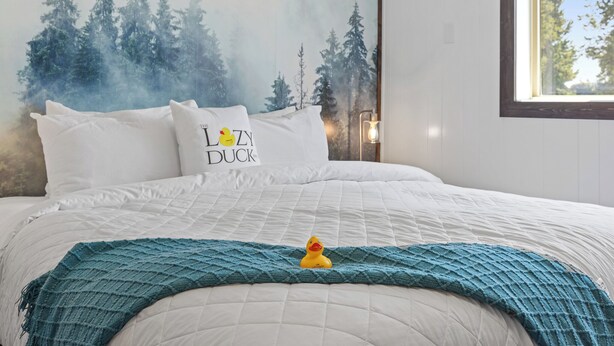 A neatly made bed with a white quilt, a teal blanket, and a pillow with a duck design. - Cozy King Suite at Lazy Duck Inn, just 1 minute from Yellowstone! #24 (West Yellowstone)