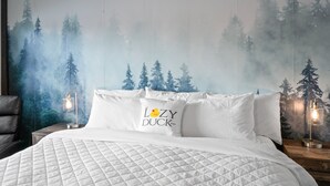 A bedroom with a bed, pillows, a nightstand, a lamp, and a wall mural of a forest. - Cozy King Suite at Lazy Duck Inn, just 1 minute from Yellowstone! #24 (West Yellowstone)