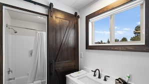 A bathroom with a shower, a toilet, a sink with a mirror, and a window with a view of trees. - Discover the Lazy Duck Inn #26 King Suite, just 1 minute from Yellowstone! (West Yellowstone)