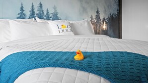 Uma cama com um cobertor azul de tricô, um pato de borracha amarelo e travesseiros com a inscrição 'The Lazy Duck'.