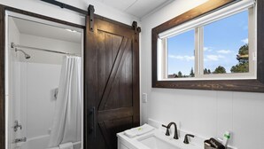 A bathroom with a shower, a toilet, a sink with a mirror, and a window with a view of trees. - Modern King Suite Lazy Duck Inn #29, just 1 minute from Yellowstone! (West Yellowstone)