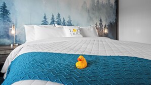 A neatly made bed with a blue knitted blanket, a white pillow with "LOZ DUCK" text, and a yellow rubber duck. - Modern King Suite Lazy Duck Inn #29, just 1 minute from Yellowstone! (West Yellowstone)