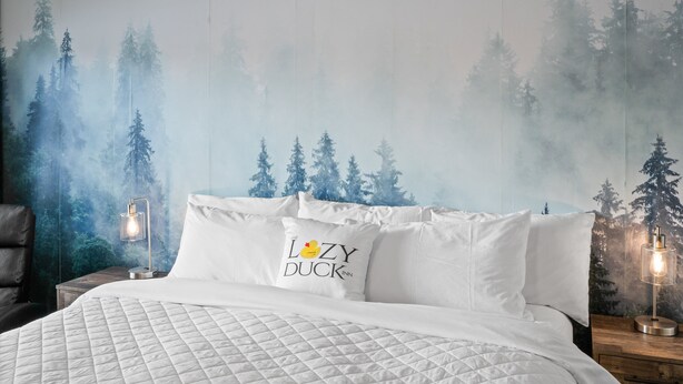 A bedroom with a bed, white bedding, a pillow with a duck design, and a wall mural of a forest. - Modern King Suite Lazy Duck Inn #29, just 1 minute from Yellowstone! (West Yellowstone)