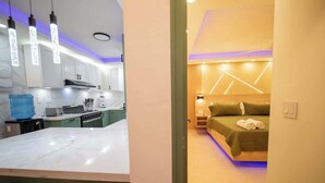 Una cocina moderna con encimera de mármol y zona de comedor. Se ve una cama con colcha verde y cabecero de madera.