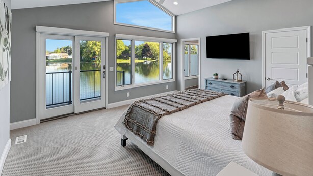 A bedroom with a large bed, a ceiling fan, a TV, and a view of a lake through large windows.