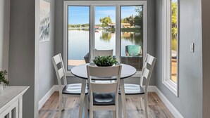 A dining area with a round table and four chairs, large windows with a view of a lake and boats, and a potted plant on the table.