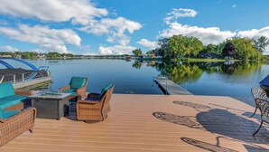 A deck with wicker furniture and a table, overlooking a calm lake with boats and houses in the background.