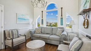 A spacious living room with a grey sofa, armchairs, and a round coffee table. Large windows offer a view of the outside. - 7223- Pelican Watch - Oceanfront Salvo | Pool & Hot Tub | Sleeps 10 (Salvo)