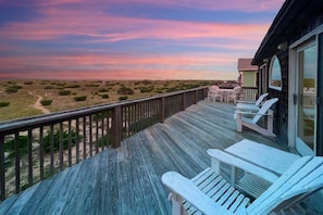 Um deck de madeira com cadeiras Adirondack brancas, com vista para uma praia com vegetação rasteira.
