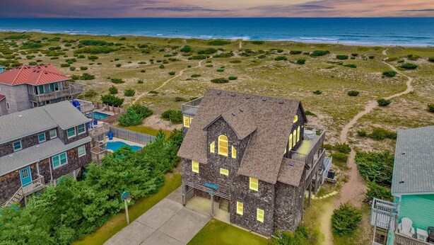 A coastal property with a large house featuring a prominent bay window, a swimming pool, and a view of the ocean. - 7223- Pelican Watch - Oceanfront Salvo | Pool & Hot Tub | Sleeps 10 (Salvo)