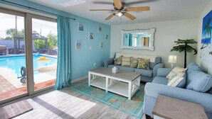 Un salon avec vue sur la piscine, un ventilateur de plafond, un canapé de couleur claire et une table basse.