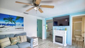 Un salon doté d’un ventilateur de plafond, d’une cheminée, d’un téléviseur et d’un tableau représentant une scène de plage.