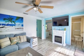 Ein Wohnzimmer mit Deckenventilator, Kamin, Fernseher und einem Strandbild.