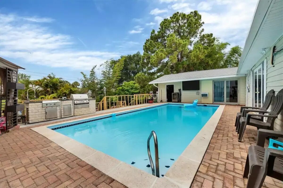 A swimming pool with a built-in barbecue and grill area, surrounded by a brick patio and lounge chairs.