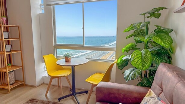 A room with a large window overlooking the sea, a yellow chair, a small table, and a potted plant. - Exclusive Panoramic Ocean View (1805) (Callao)