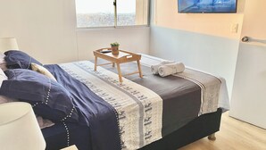 A neatly made bed with a patterned bedspread, a small wooden table with a plant, and a pair of towels. A television is mounted on the wall displaying a snowy landscape. - Exclusive Panoramic Ocean View (1805) (Callao)