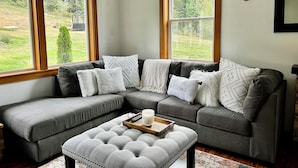 A corner living room with a grey sectional sofa, multiple cushions, a white ottoman, and a remote control on a tray. - Wildcat Cottage  (Port Angeles)
