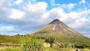 A mountain with a conical shape, surrounded by greenery and a partly cloudy sky. - Hospedaje Peña del Arenal (La Fortuna)