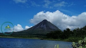 A mountain with a lake in the foreground and a logo in the bottom left corner. - Hospedaje Peña del Arenal (La Fortuna)